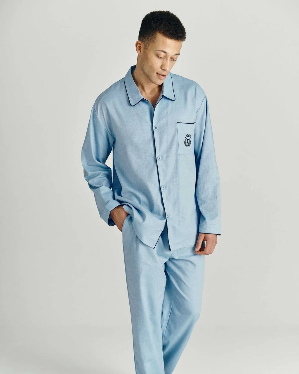 Maison Sarava light blue cotton pajama set with black piping and embroidered crest on pocket. Model wearing button-down shirt and relaxed-fit pants.