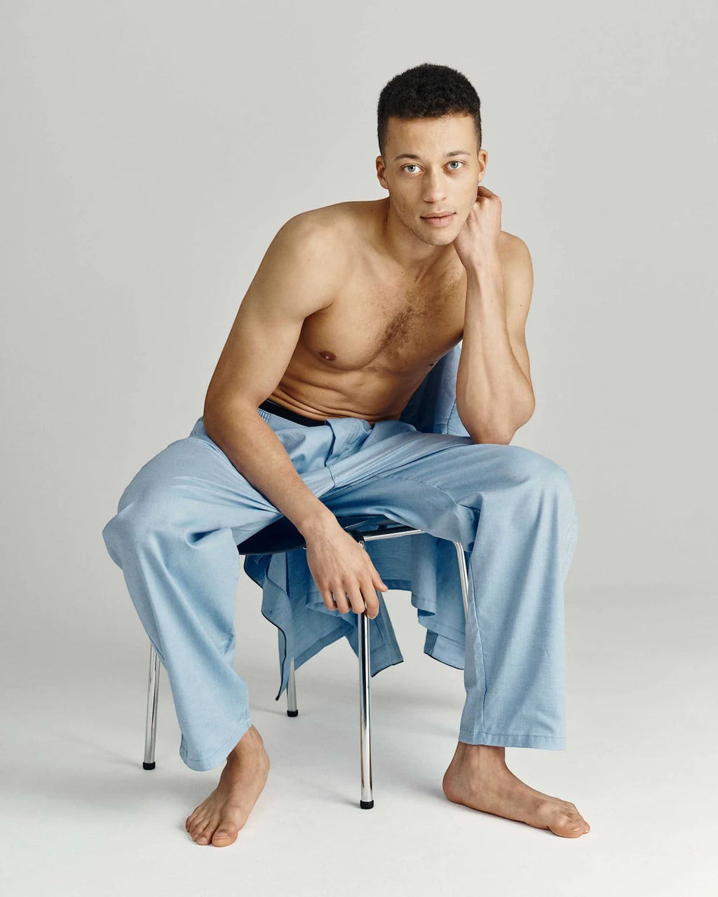 Maison Sarava light blue cotton pajama set with black piping. Model seated, wearing pajama pants and draped shirt.