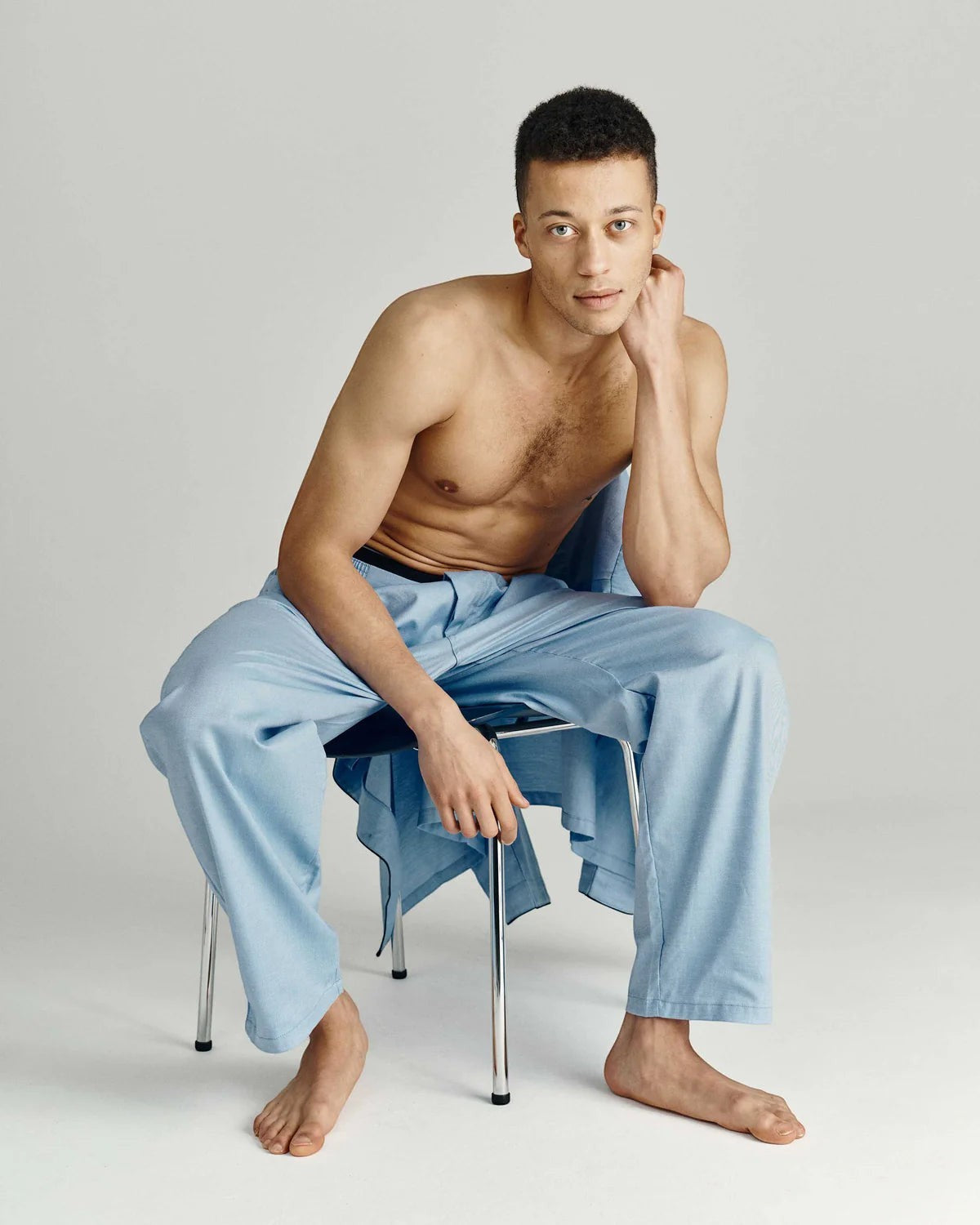 Maison Sarava light blue cotton pajama set with black piping. Model seated, wearing pajama pants and draped shirt.